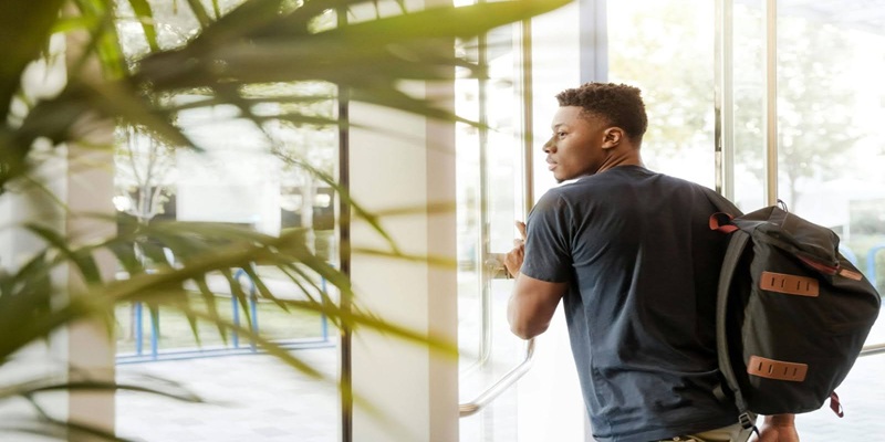 College student Looking Outside Window Carrying Black and Brown Backpack While Holding His Hand on Window
